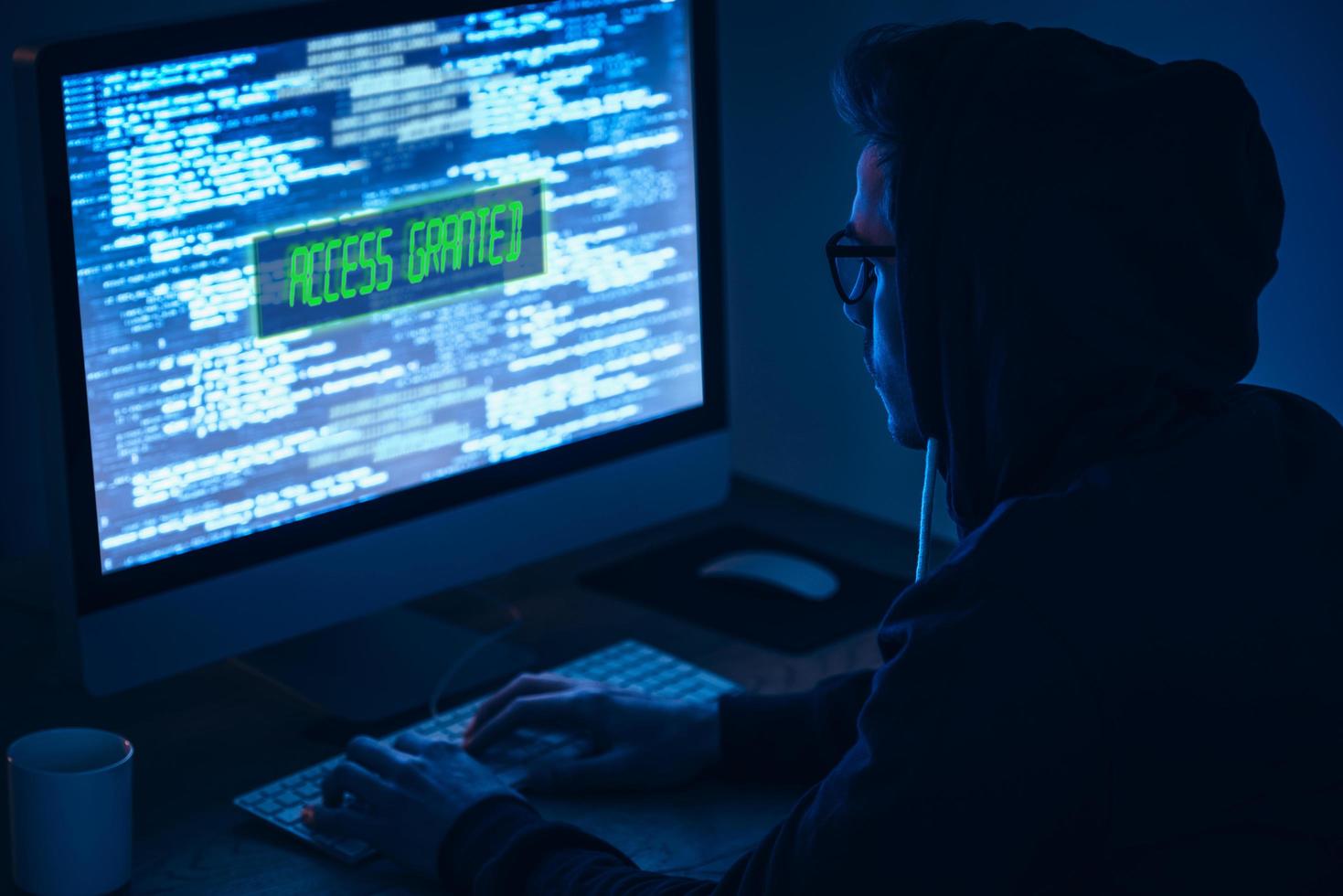 Computer hacker. Rear view of young man typing and looking at computer monitor while sitting at the table in dark room photo