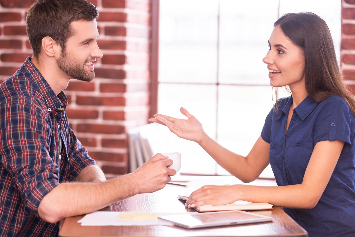 Colleagues on coffee break. Side view of cheerful young man and woman sitting at the table and chatting while man holding cup of hot drink photo