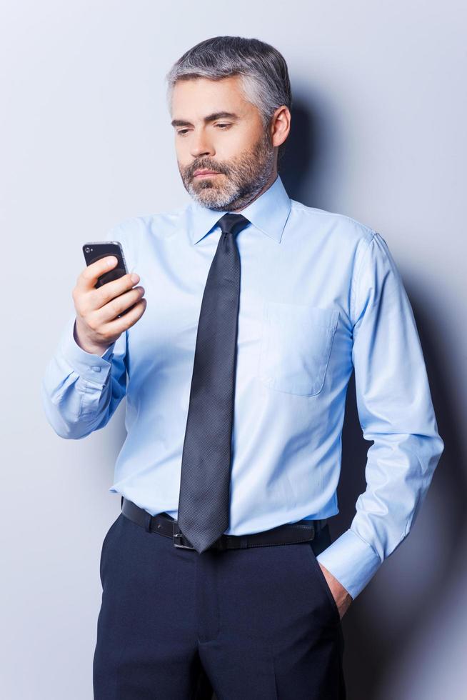 revisando mensajes de negocios. hombre maduro seguro de sí mismo con camisa y corbata sosteniendo un teléfono móvil y mirándolo mientras se enfrenta a un fondo gris foto