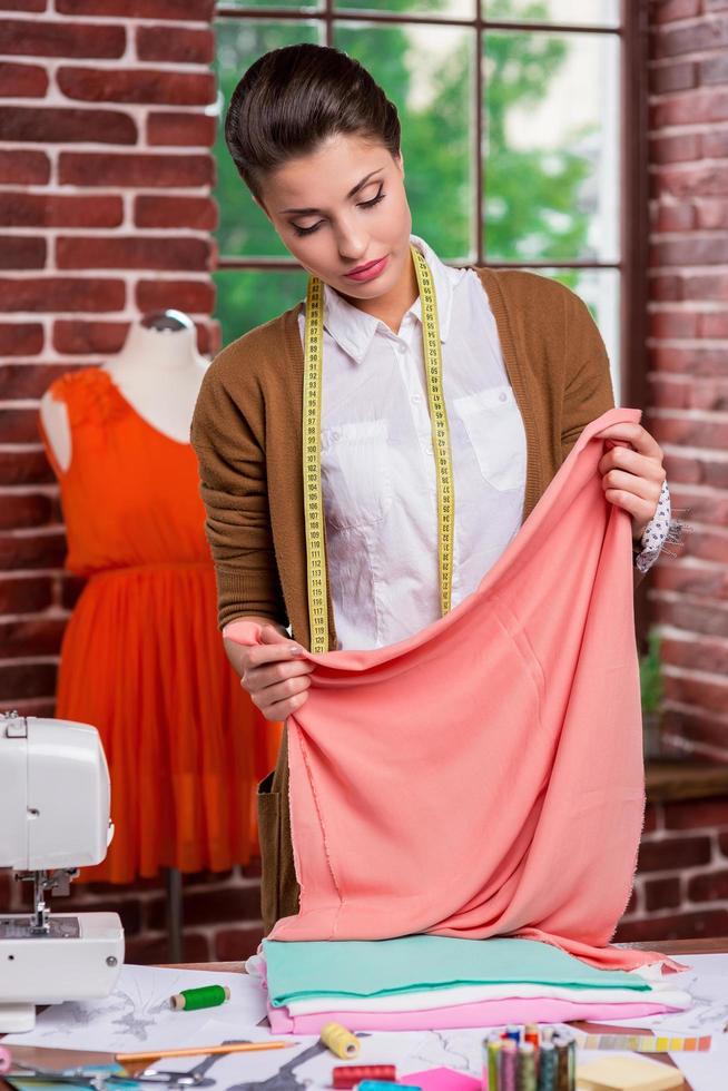 Examining textile before sewing. Beautiful female fashion designer holding a stack of multi colored textile while standing near her working place and with mannequin in the background photo