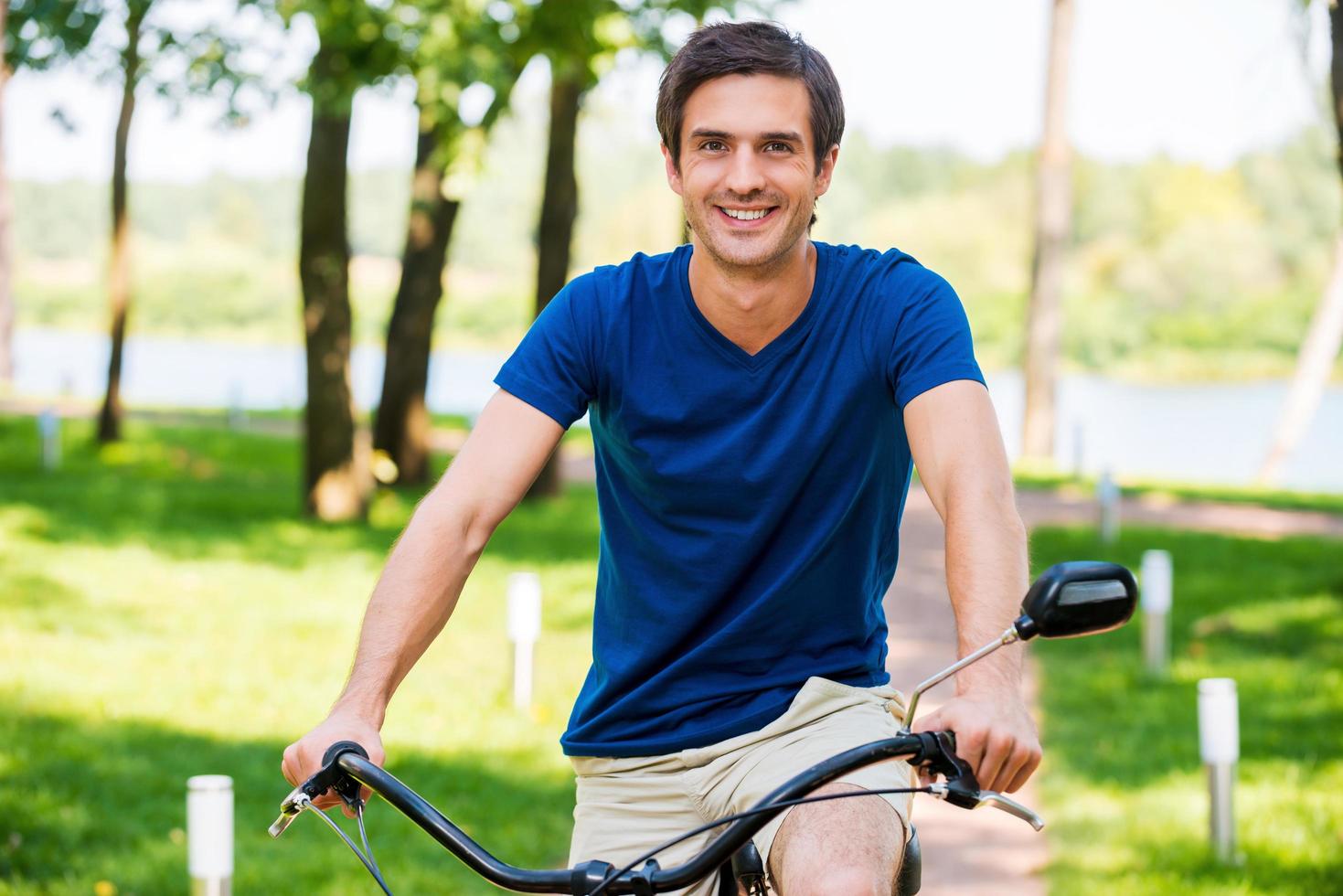 Enjoying his park ride. Cheerful young man riding bicycle in park and smiling at camera photo