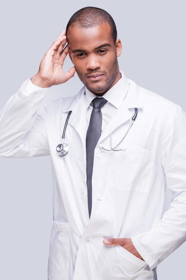 Feeling uncertain. Thoughtful African doctor touching head with hand and looking away while standing against grey background photo