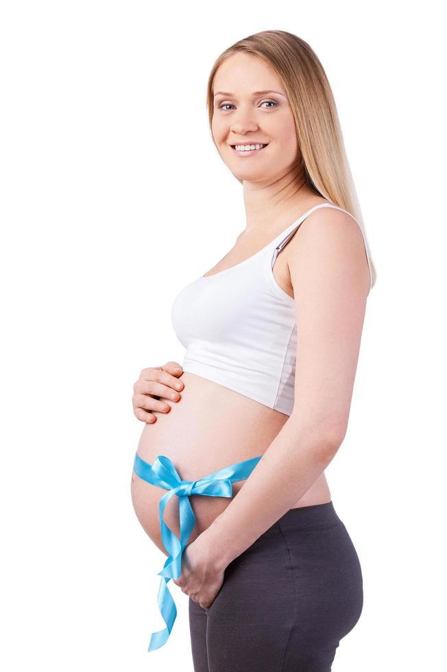 Expecting a baby boy. Side view of happy pregnant woman with blue ribbon on her belly looking at camera and smiling while standing isolated on white photo