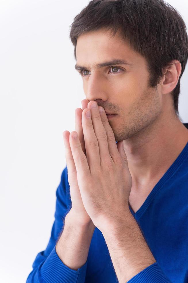 Man praying. Side view of thoughtful young man in white shirt holding hands clasped near face and looking away while standing against grey background photo
