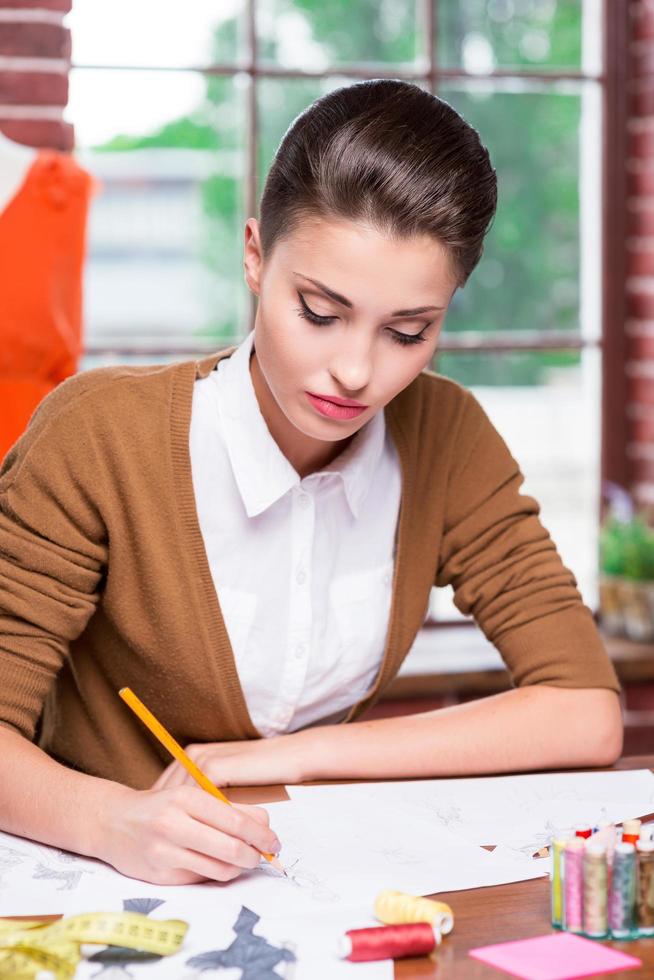 Working on creative fashion look. Beautiful young woman drawing fashion sketch while sitting at her working place photo