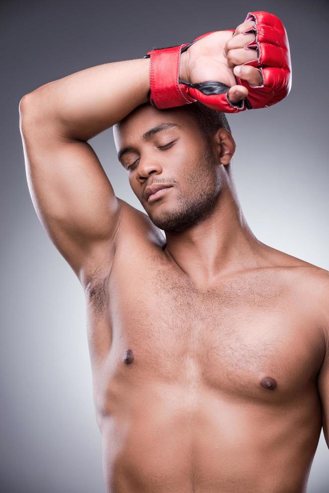 Tired after fight. Young shirtless African man in boxing gloves touching his forehead and keeping eyes closed while standing against grey background photo