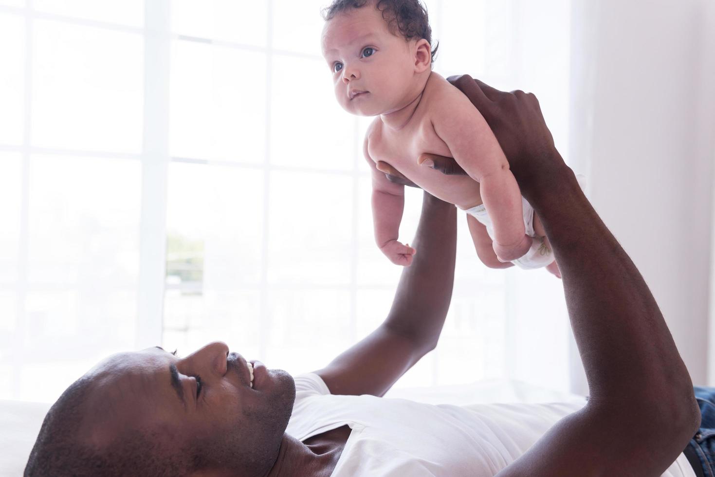 padre orgulloso y feliz. vista lateral de un joven africano feliz levantando a su pequeño bebé y sonriendo mientras está acostado en la cama foto