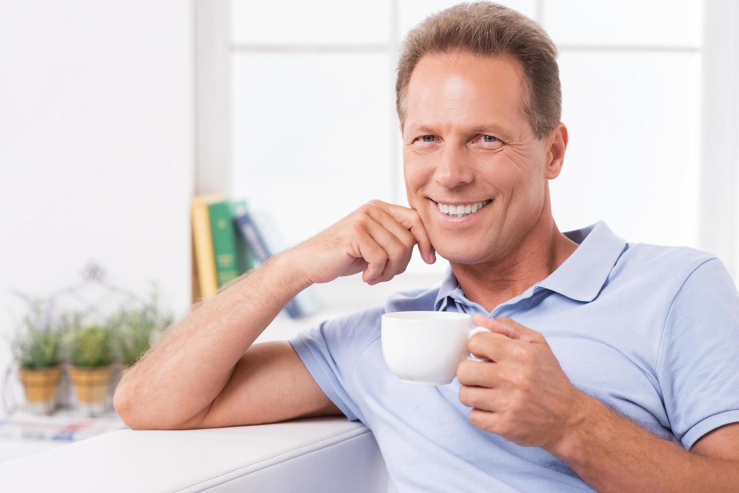 relajarse con café recién hecho y las últimas noticias. hombre maduro alegre bebiendo café y leyendo el periódico mientras se sienta en el sofá en casa foto