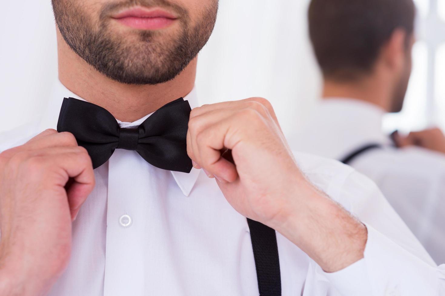 Adjusting his bow tie. Close-up of young man in white shirt adjusting his bow tie while standing against mirror photo