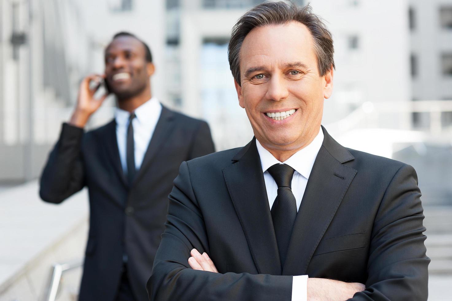 experto en el mundo empresarial. hombre mayor confiado en ropa formal con los brazos cruzados y sonriendo mientras el hombre africano habla por teléfono móvil en el fondo foto