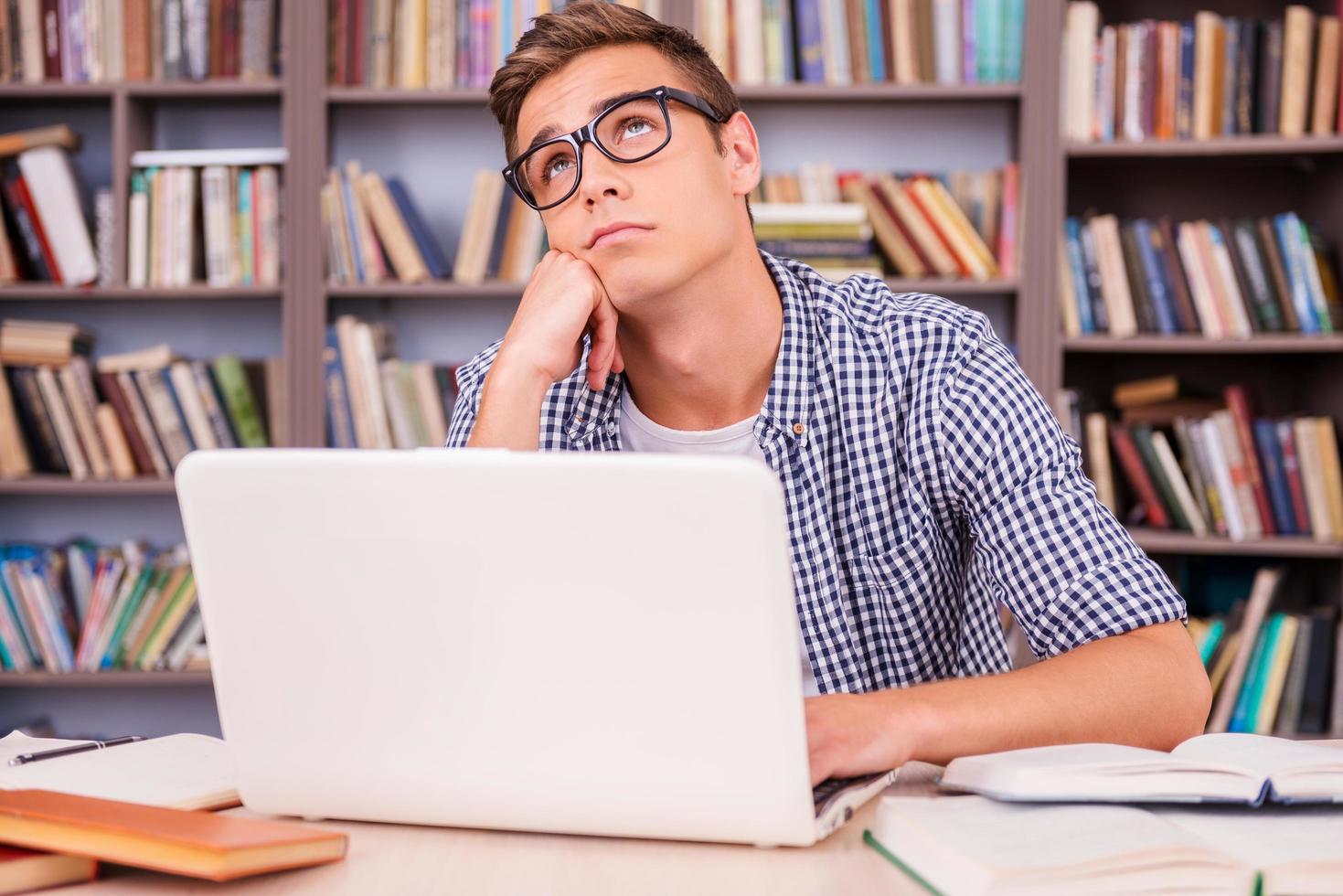 Day dreaming in library. Bored young man leaning his face on hand and looking away while sitting at the desk and in font of bookshelf photo