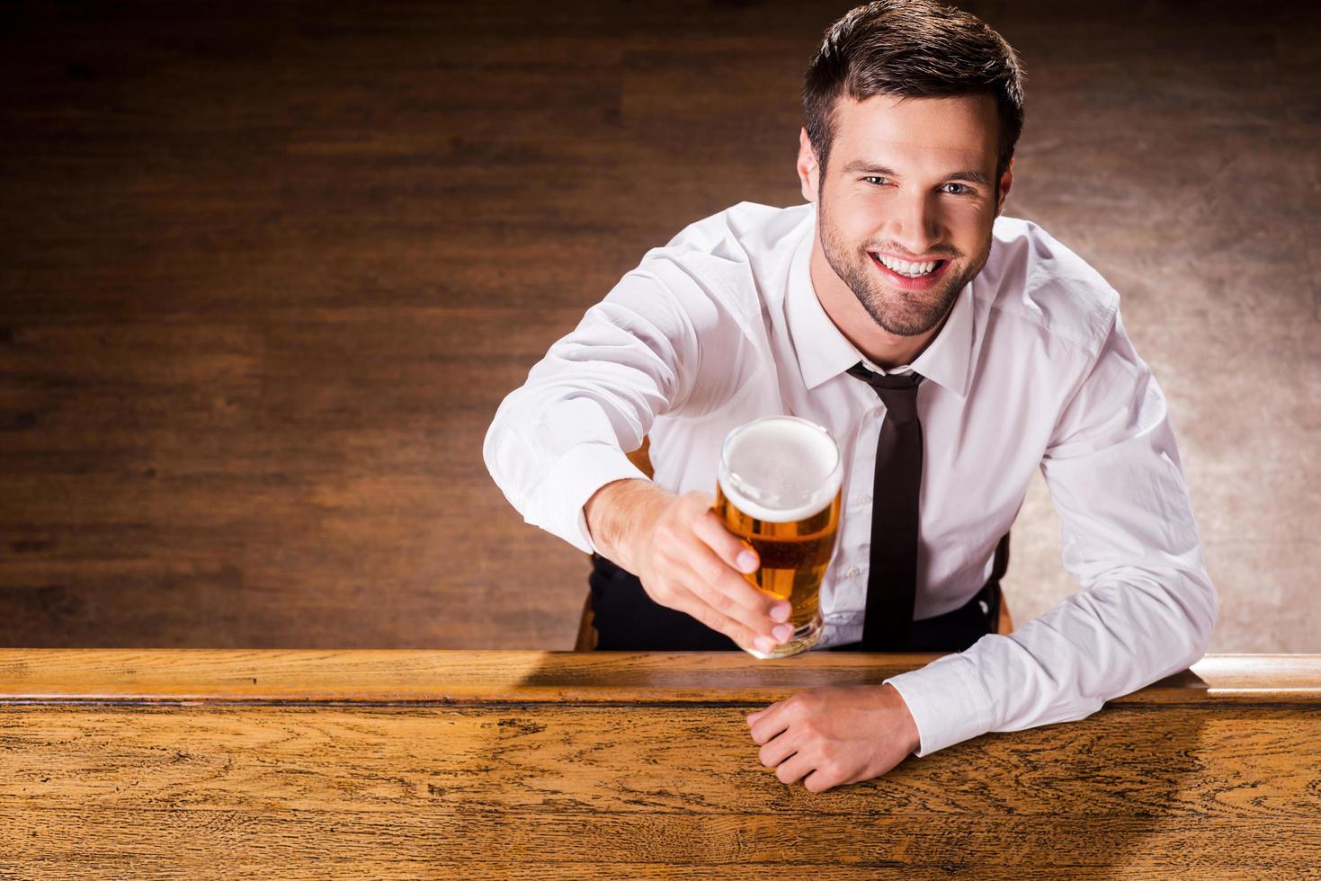 relajante con un vaso de cerveza fresca. vista superior de un apuesto joven con camisa y corbata sosteniendo un vaso con cerveza y sonriendo mientras se sienta en el mostrador del bar foto