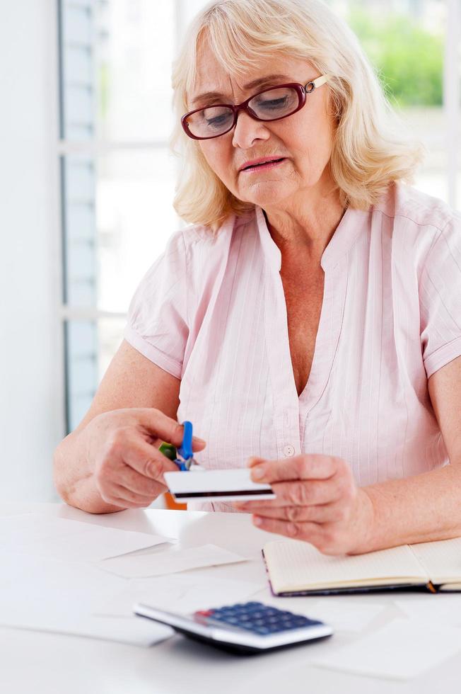 recortando su presupuesto. anciana concentrada cortando su tarjeta de crédito con tijeras mientras se sienta en la mesa foto