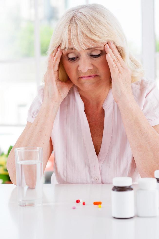 Feeling headache. Depressed senior woman holding head in hands and looking at the pills laying on the table photo