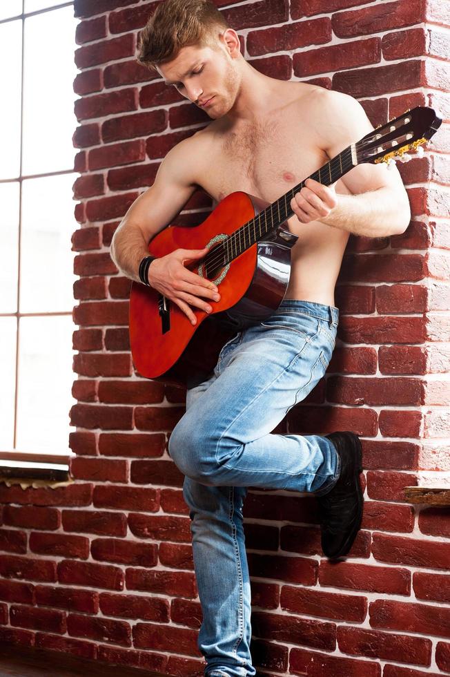 Playing guitar. Handsome young shirtless man playing acoustic guitar while leaning at the brick wall photo