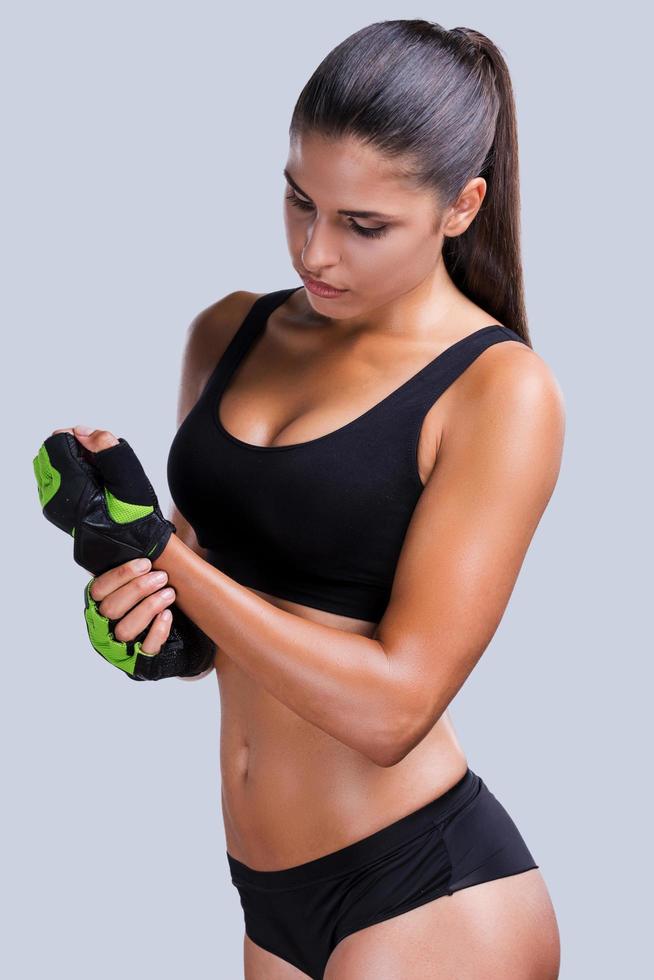 Preparing to sports training. Beautiful young sporty woman with perfect body adjusting her sports gloves while standing against grey background photo