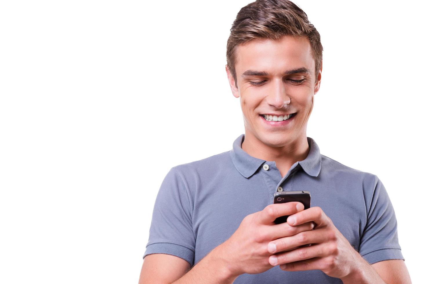 Sending message to friend. Happy young man holding mobile phone and looking at it with smile while standing isolated on white background photo