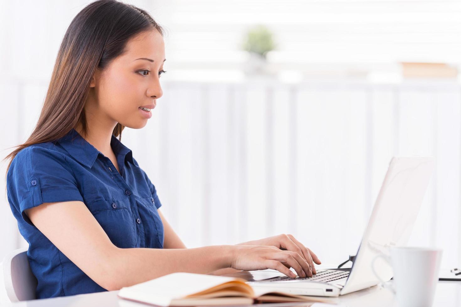 Surfing the latest news online. Side view of beautiful young Asian woman working on laptop and smiling while sitting at her working place photo