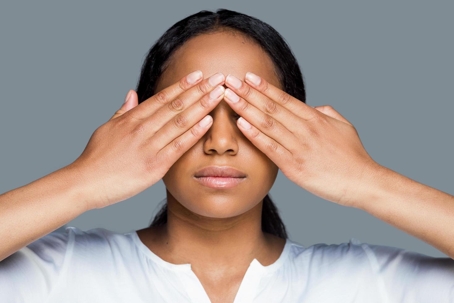 I see nothing. Beautiful young African woman covering eyes with hands while standing against grey background photo