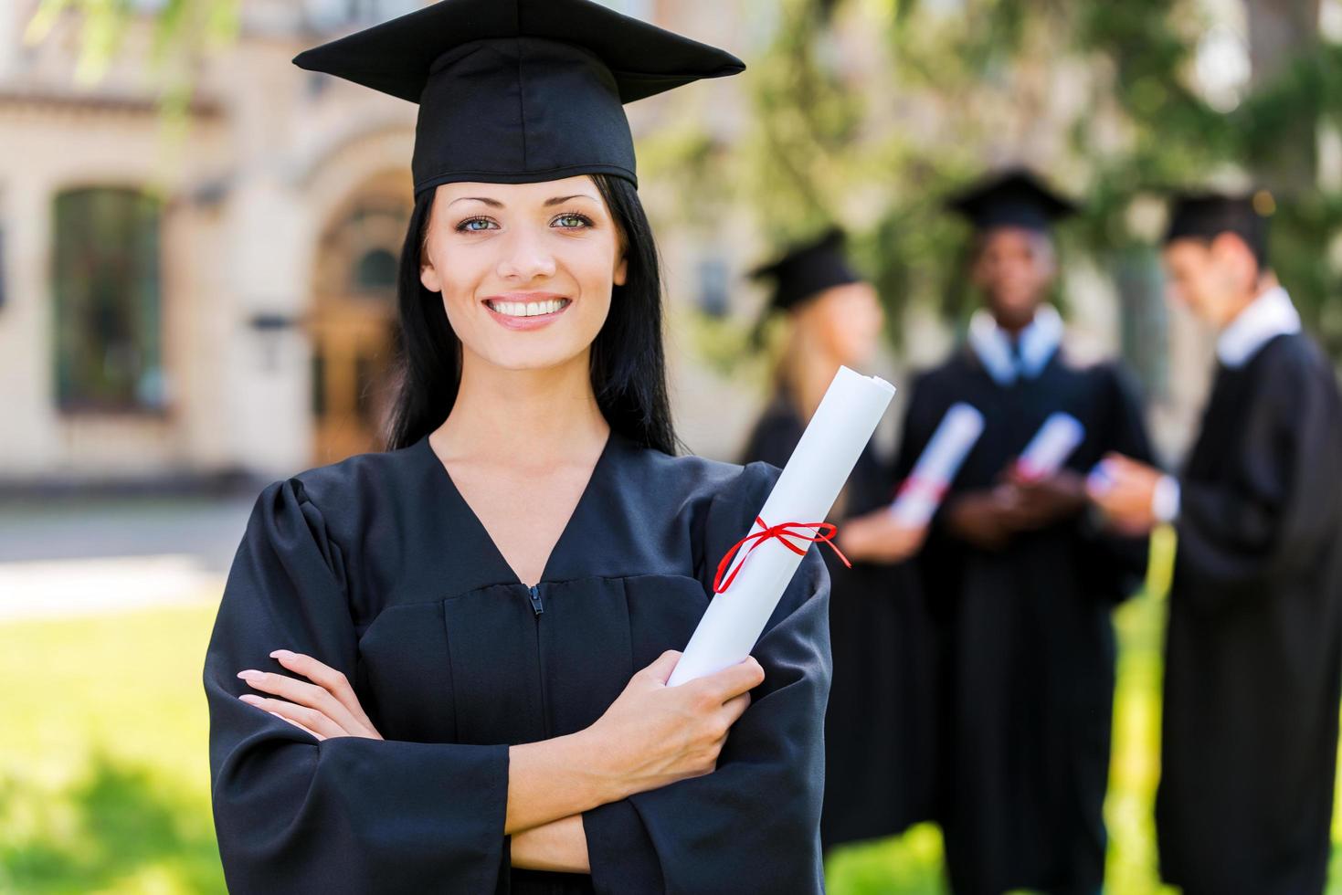 Happy to be graduated. Happy young woman in graduation gowns holding diploma and smiling while her friends standing in the background photo