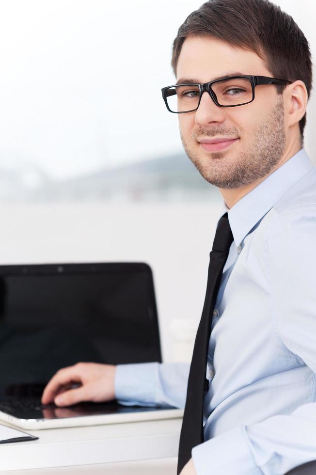 Confident businessman. Rear view of cheerful young man in shirt and tie looking over shoulder while sitting at his working place photo