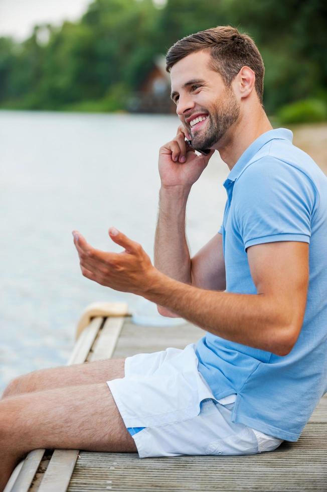 Good talk with friend. Side view of handsome young man talking on the mobile phone and smiling while sitting at the riverbank photo