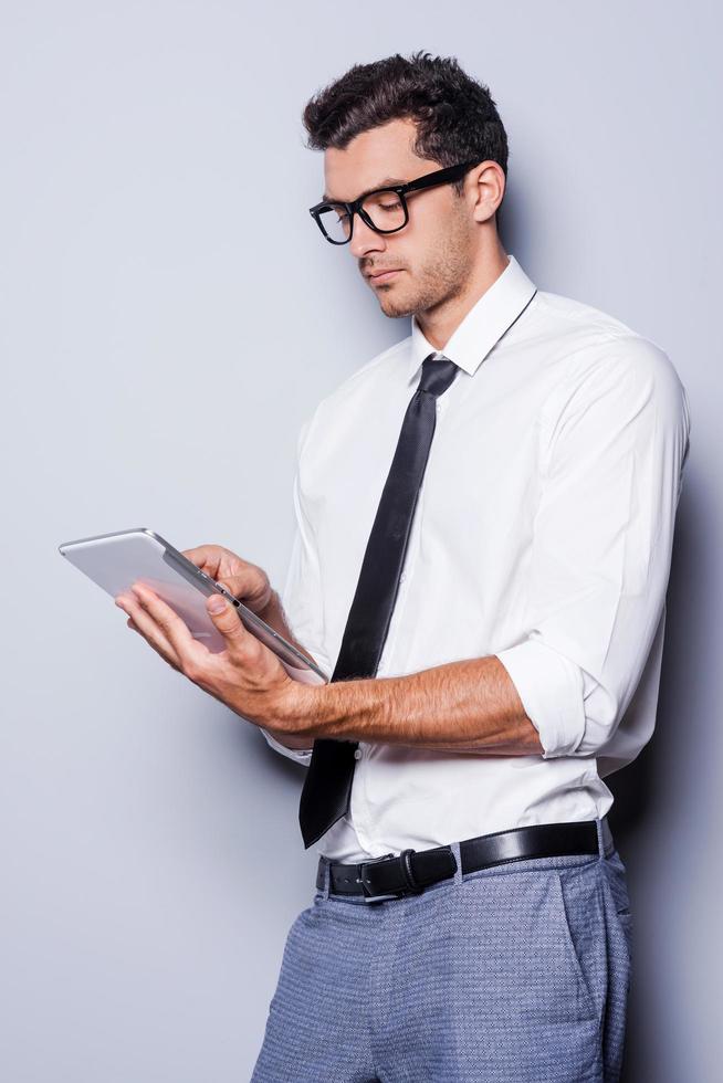 Working on digital tablet. Handsome young man in shirt and tie working on digital tablet while standing against grey background photo