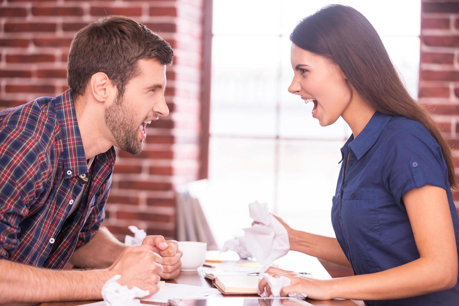 Battle of the sexes. Side view image of angry man and woman sitting face to face at the office table and shouting at each other photo