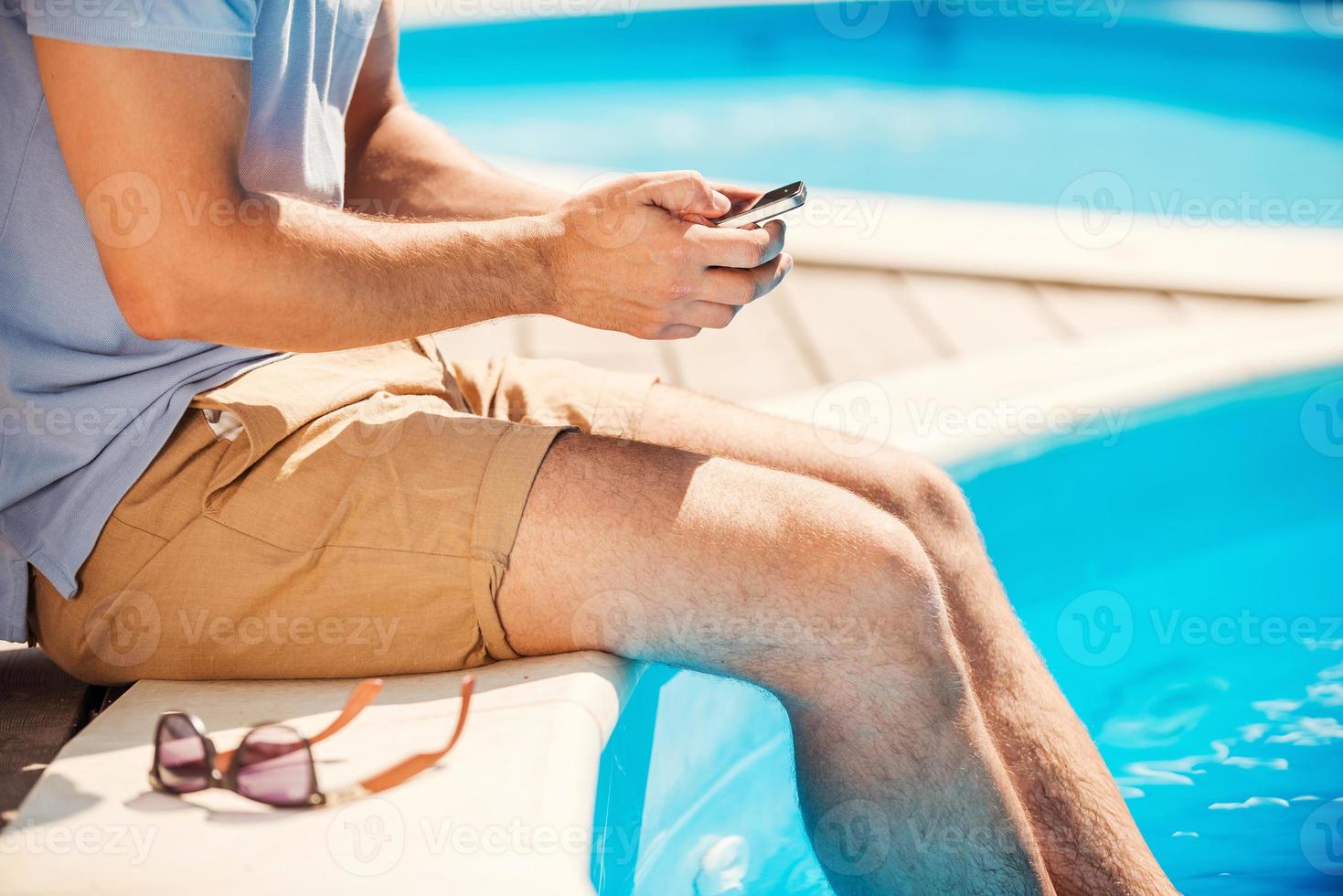 Chatting with friends. Close-up side view of man sitting poolside and typing something on mobile phone photo