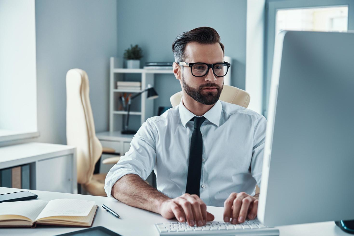 joven ocupado con camisa y corbata trabajando en la computadora mientras está sentado en la oficina foto