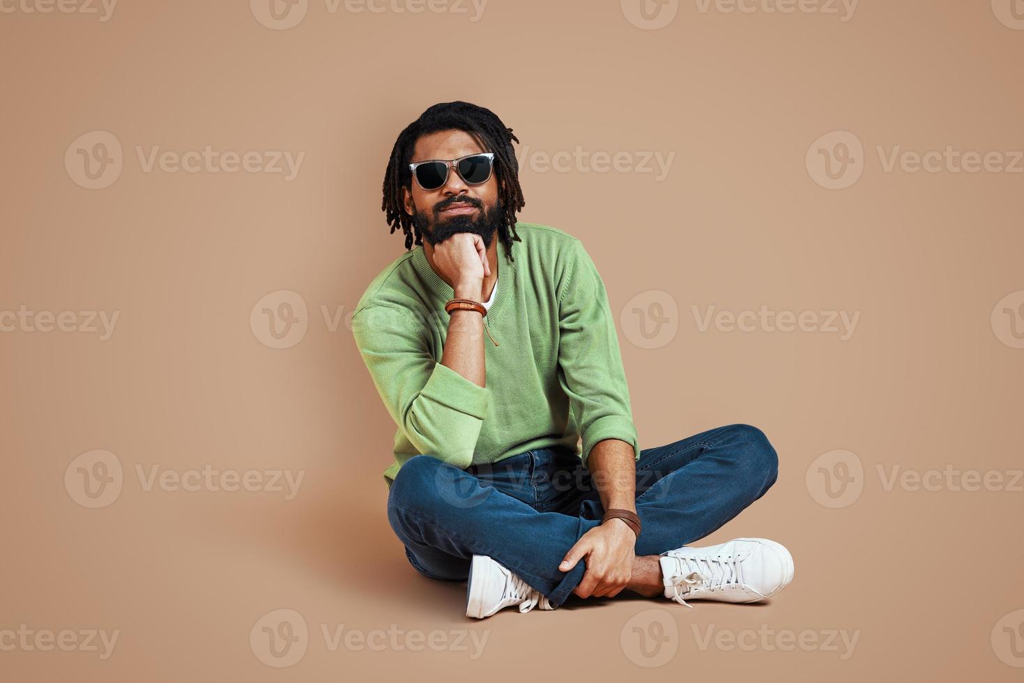 joven africano de moda con ropa informal mirando la cámara y sonriendo mientras se sienta en un fondo marrón foto