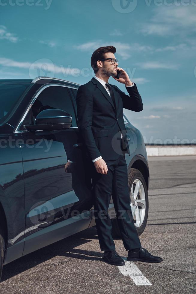 hablando de negocios. toda la longitud de un apuesto joven hombre de negocios hablando por teléfono mientras está parado cerca de su auto al aire libre foto