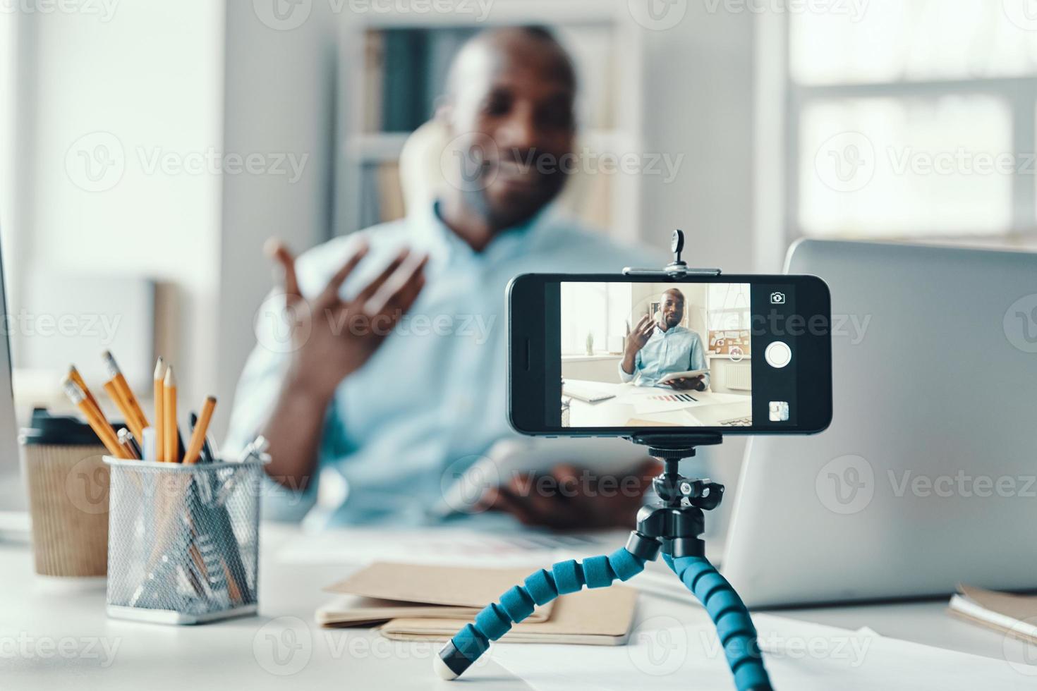 Handsome young African man in shirt telling something and smiling while making social media video photo