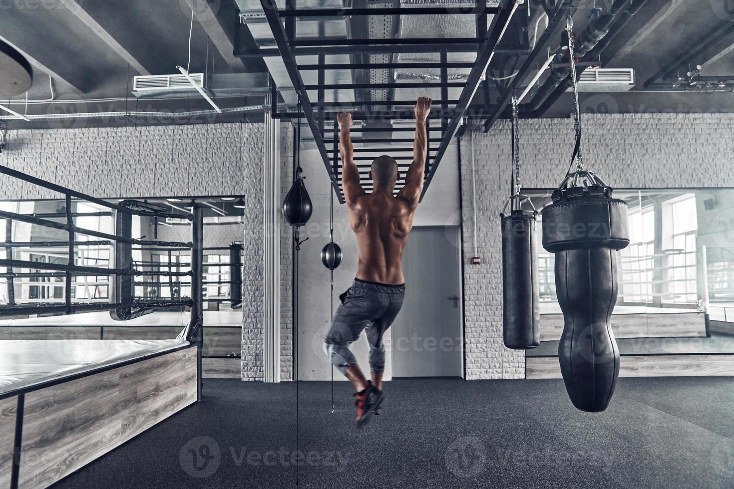 Towards the healthier lifestyle. Full length rear view of shirtless young African man doing pull-ups while exercising in the gym photo