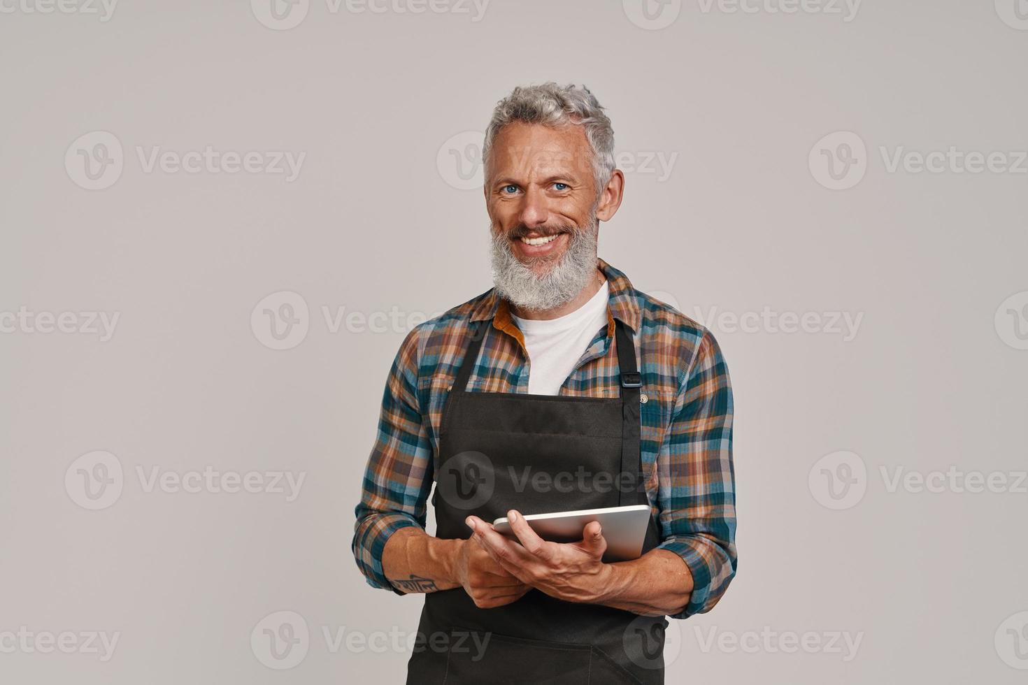 Senior man in apron smiling and using digital tablet while standing against gray background photo