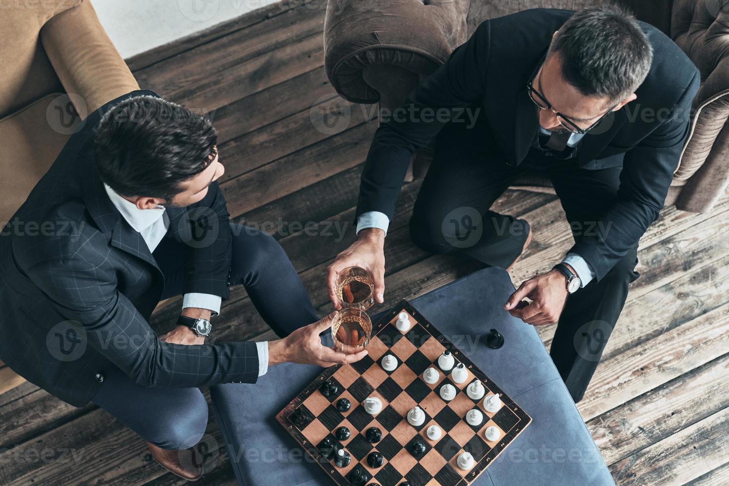 a la vista superior del juego interesante de hombres jóvenes reflexivos en trajes completos jugando al ajedrez mientras están sentados en el interior foto