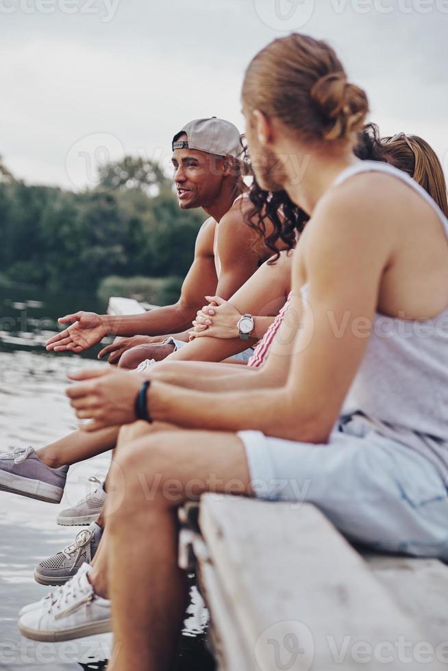 Friendly talk. Group of happy young people in casual wear smiling and talking while sitting on the pier photo