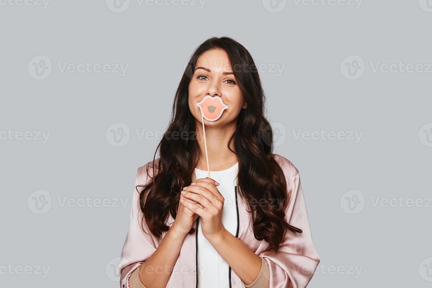 Funky style. Attractive young woman wearing costume human lips on the prop and looking at camera while standing against grey background photo