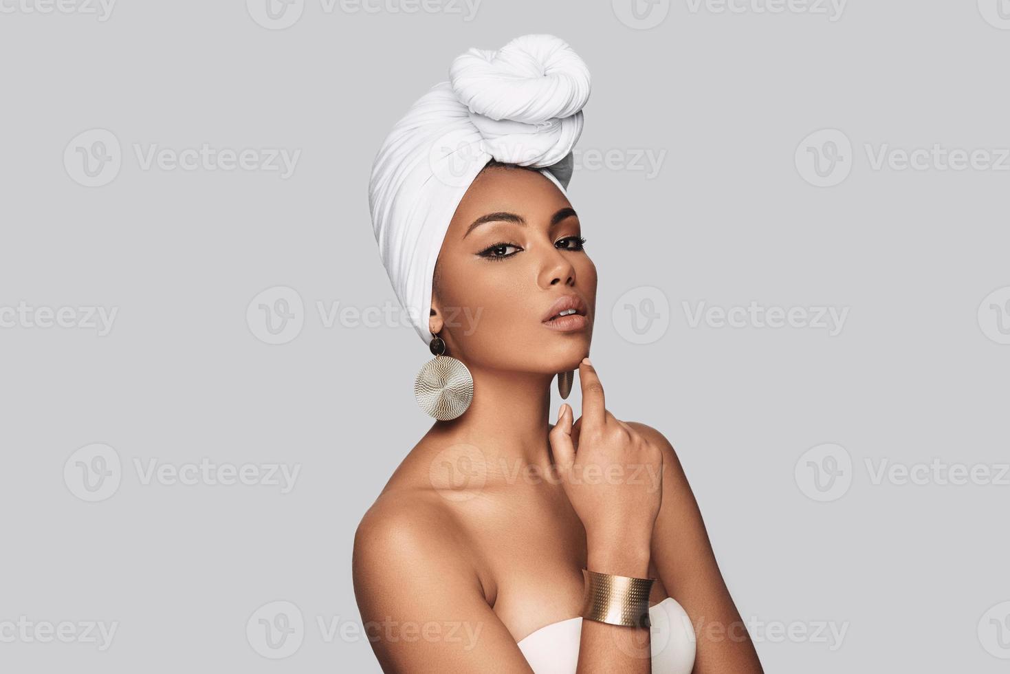 Impossible to resist her. Attractive young African woman in turban keeping hand on chin while standing against grey background photo