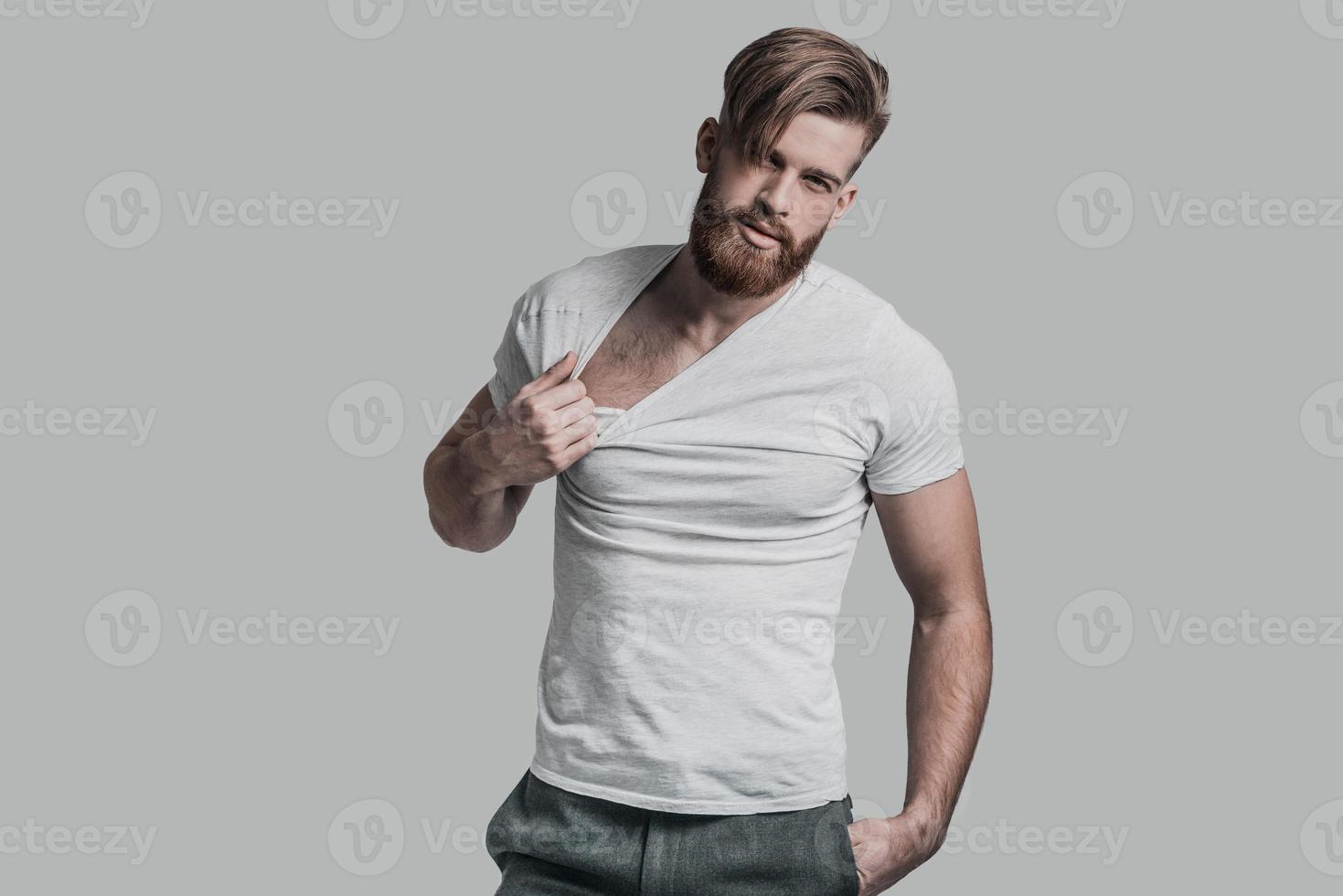 genial y guapo. joven tirando de su camiseta mientras está de pie contra el fondo gris foto