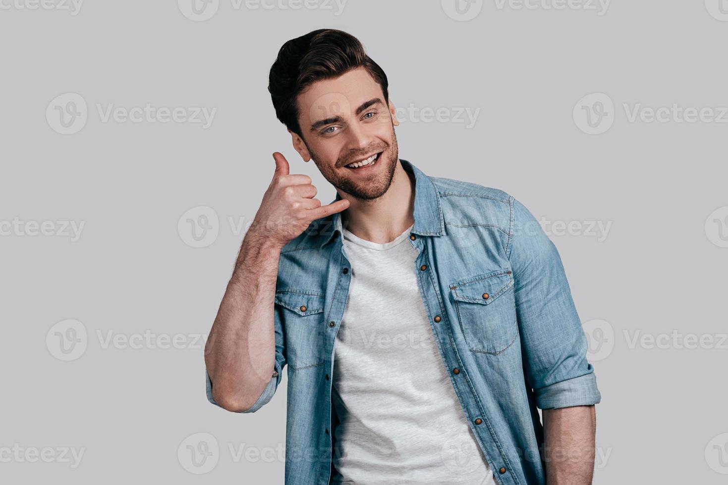 Give me a call Good looking young man in blue jeans shirt gesturing and looking at camera while standing against grey background photo