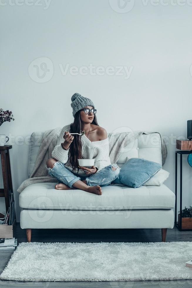 disfrutando de su comida. bella joven con ropa informal y sombrero de punto sosteniendo plato y gente y mirando hacia otro lado mientras se sienta en el sofá de casa foto