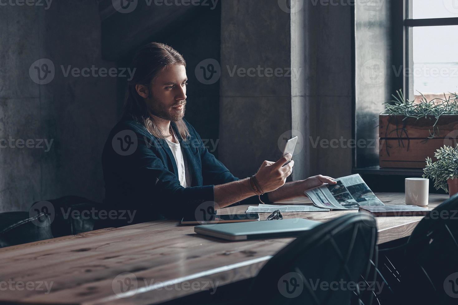 Sending message.  Handsome young man in smart casual wear holding smart phone and looking at it while sitting at his desk in office photo