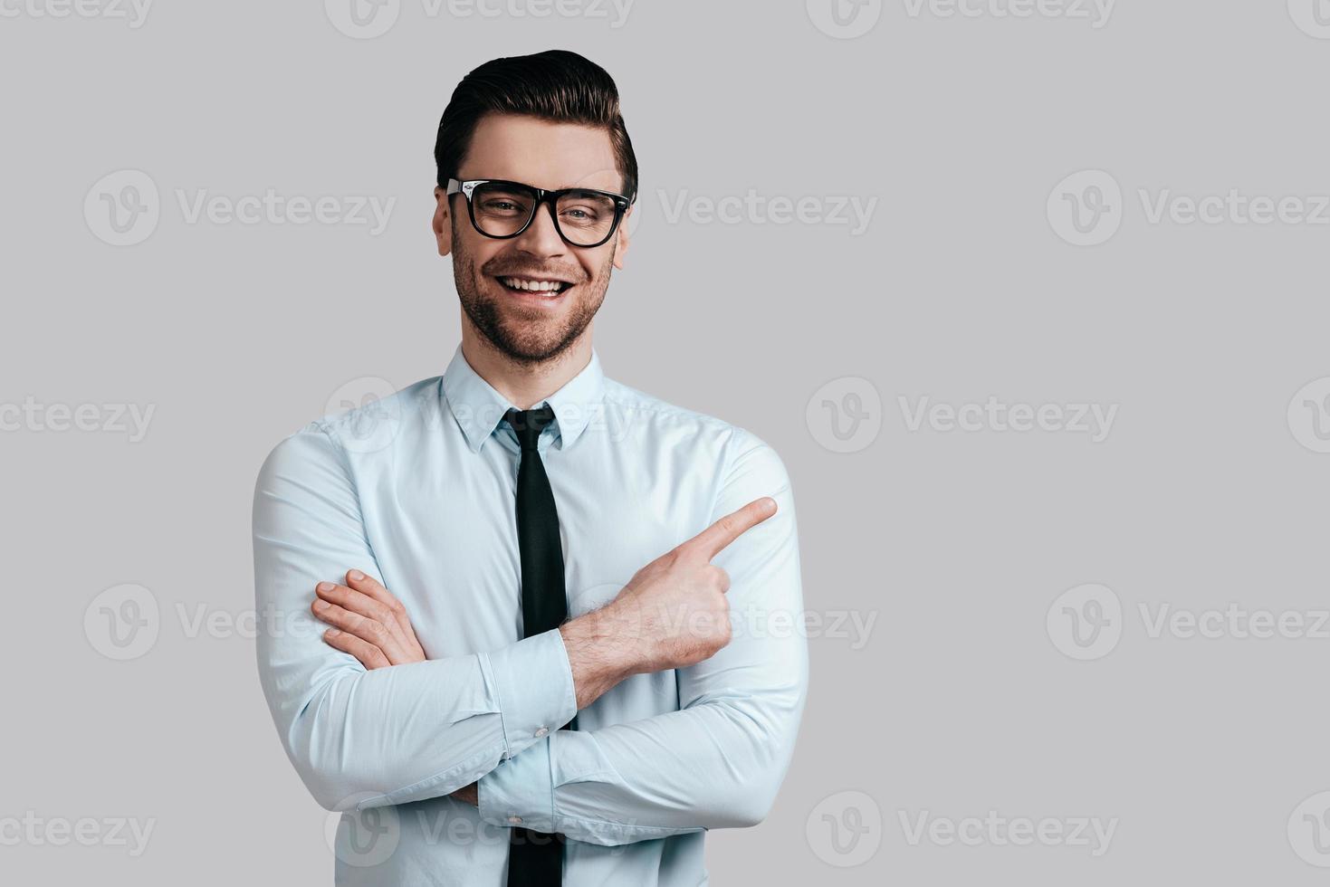 Pointing copy space. Handsome smiling man in eyeglasses keeping arms crossed and looking at camera while standing against grey background photo