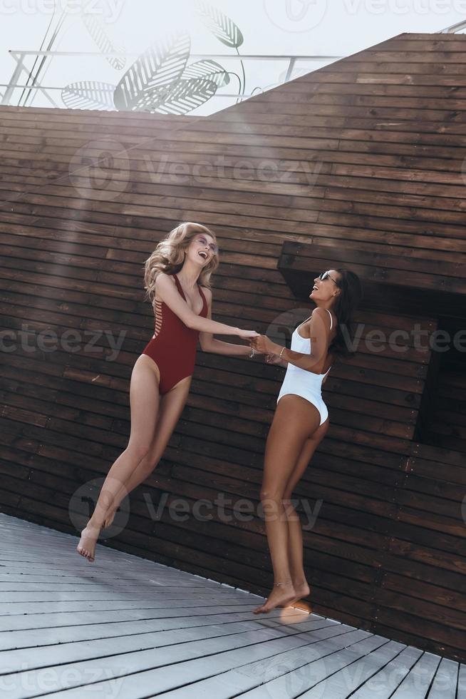 Spontaneous dance. Full length of two attractive young women in swimwear smiling while dancing against the wooden wall outdoors photo
