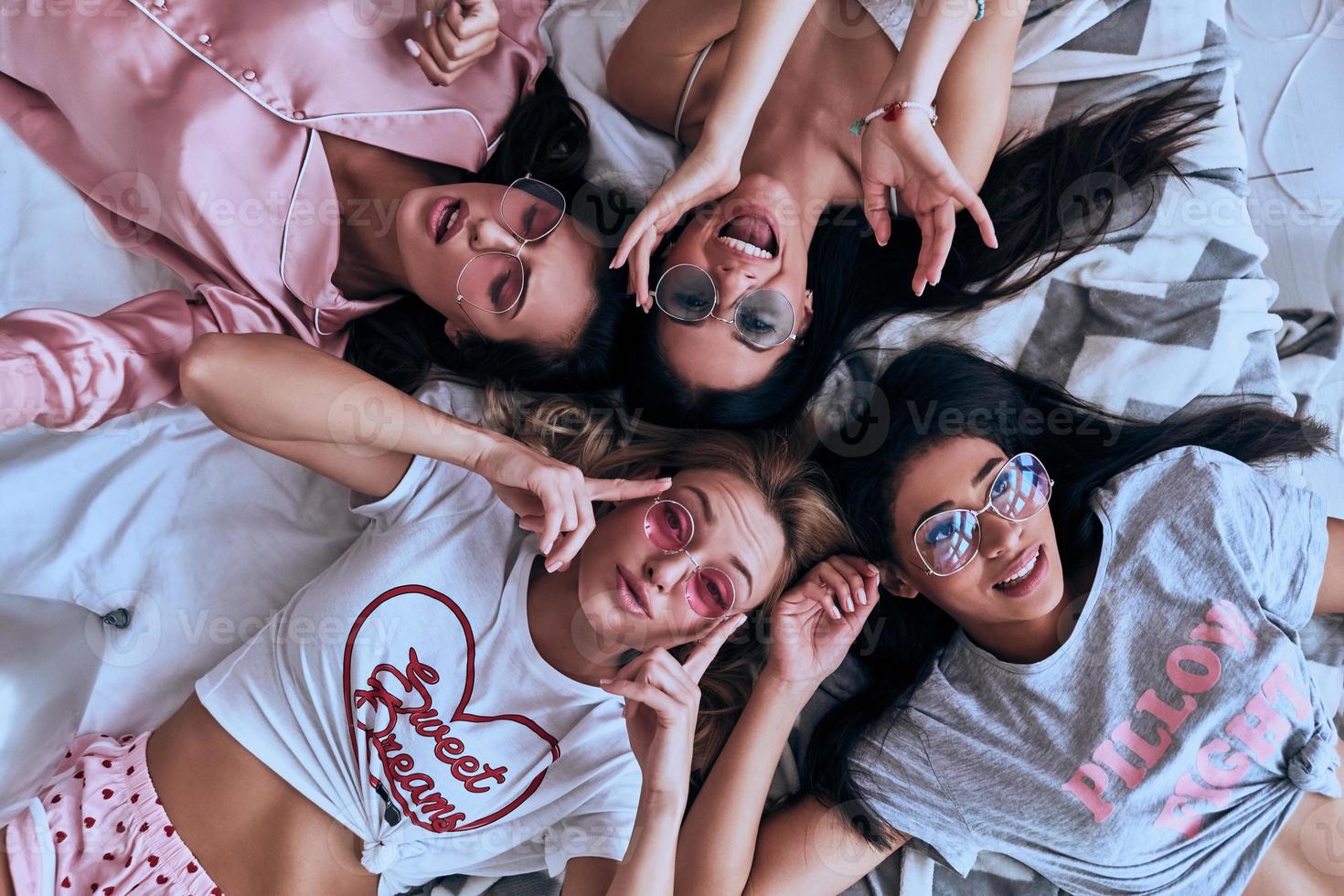 Feeling free to be happy. Top view of four playful young women in pajamas and eyewear smiling while lying on the bed at home photo