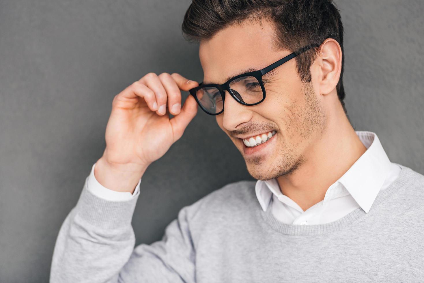 Elegance and success. Side view of confident young man adjusting his glasses and looking away with smile while standing against grey background photo