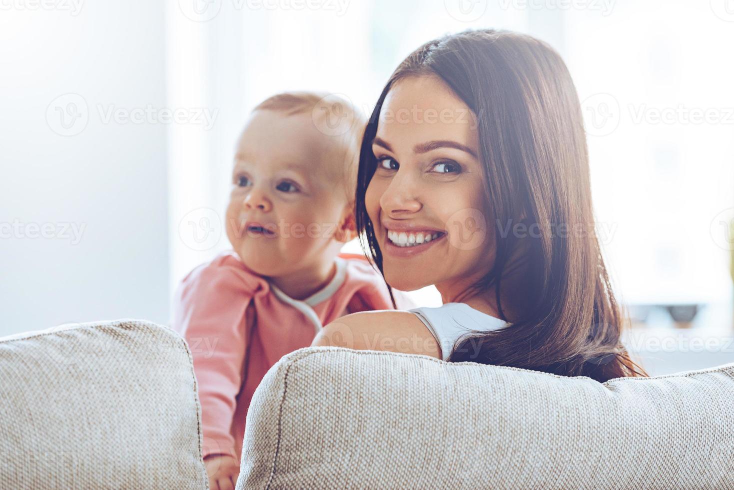 Young mommy. Rear view of cheerful beautiful young woman holding baby girl in her hands and looking at camera with smile while sitting on the couch at home photo