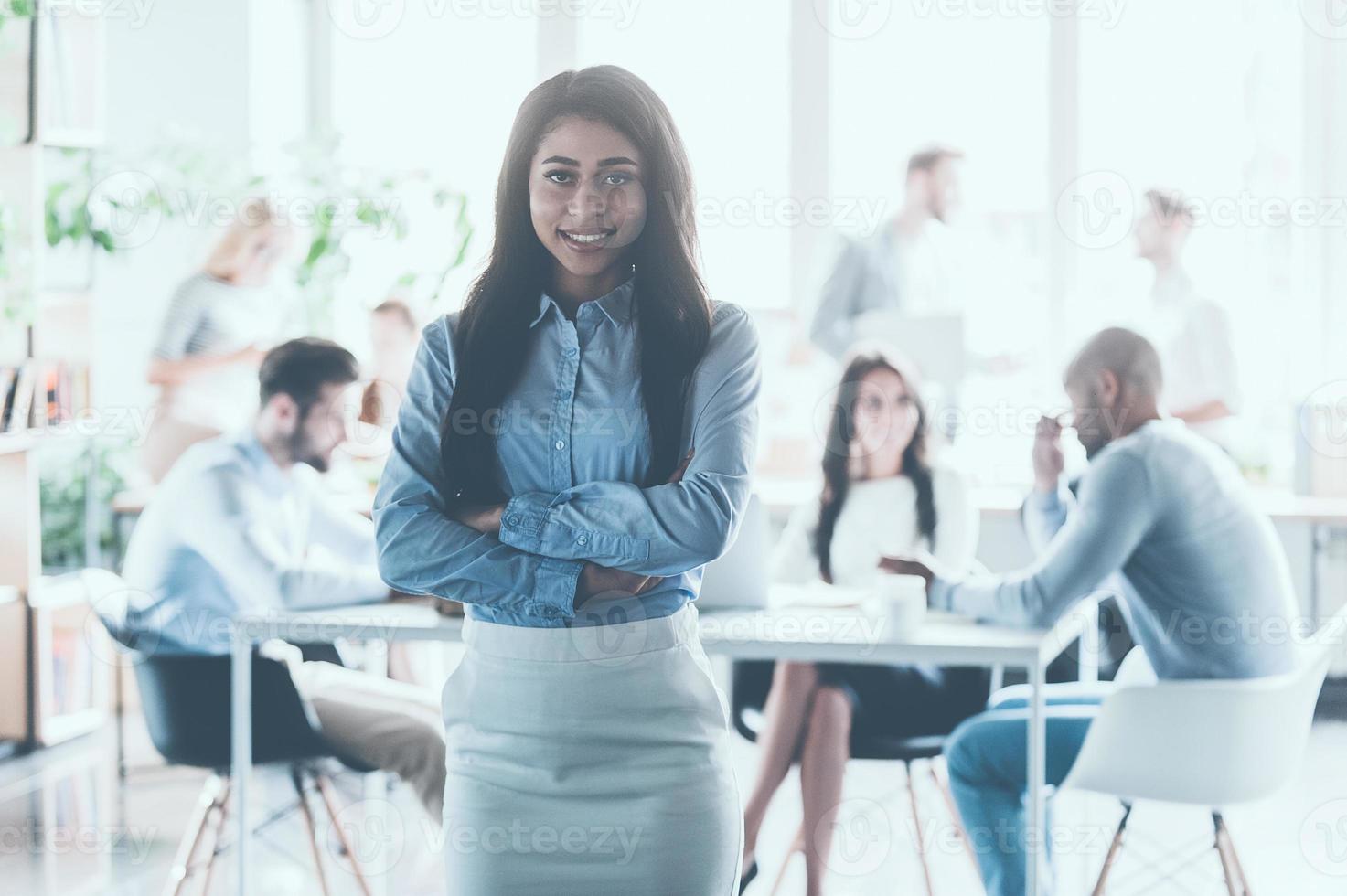 Feeling confident in her team. Beautiful young African woman keeping arms crossed and looking at camera with smile while her colleagues working in the background photo