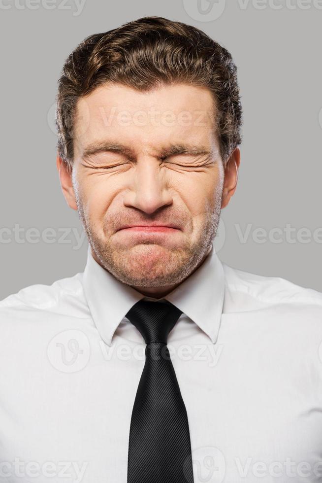 Businessman grimacing. Frustrated young man in shirt and tie keeping eyes closed and grimacing while standing against grey background photo